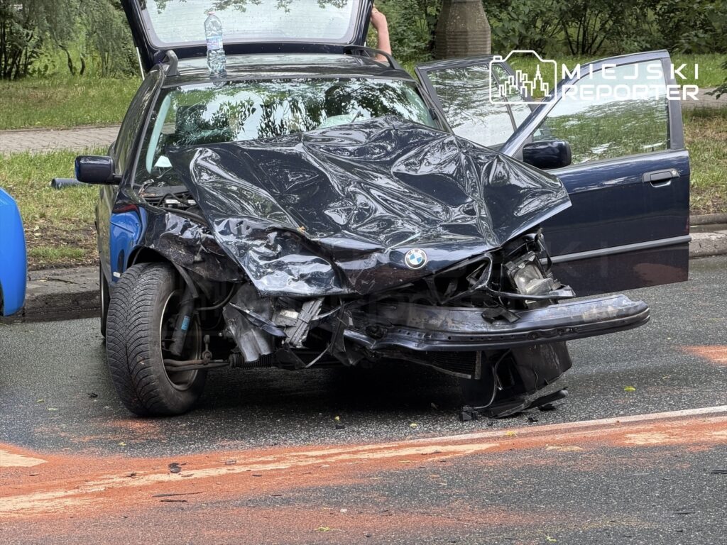 Zniszczony samochód osobowy marki BMW zaparkowany na drodze, z uszkodzoną przednią częścią i otwartymi drzwiami.