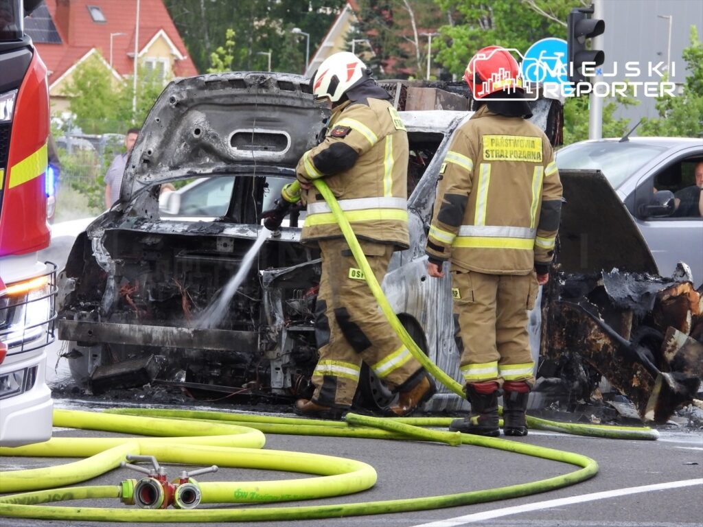 Strażacy gaszą płonący samochód osobowy na jezdni, obok leżą węże gaśnicze, w tle widać budynki mieszkalne.