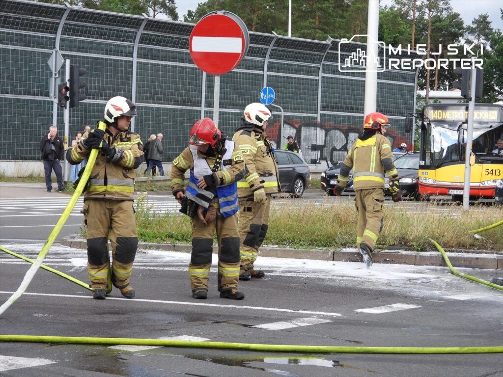 Czterech strażaków w mundurach operacyjnych używa węża gaśniczego na skrzyżowaniu, obok przejeżdżających samochodów.