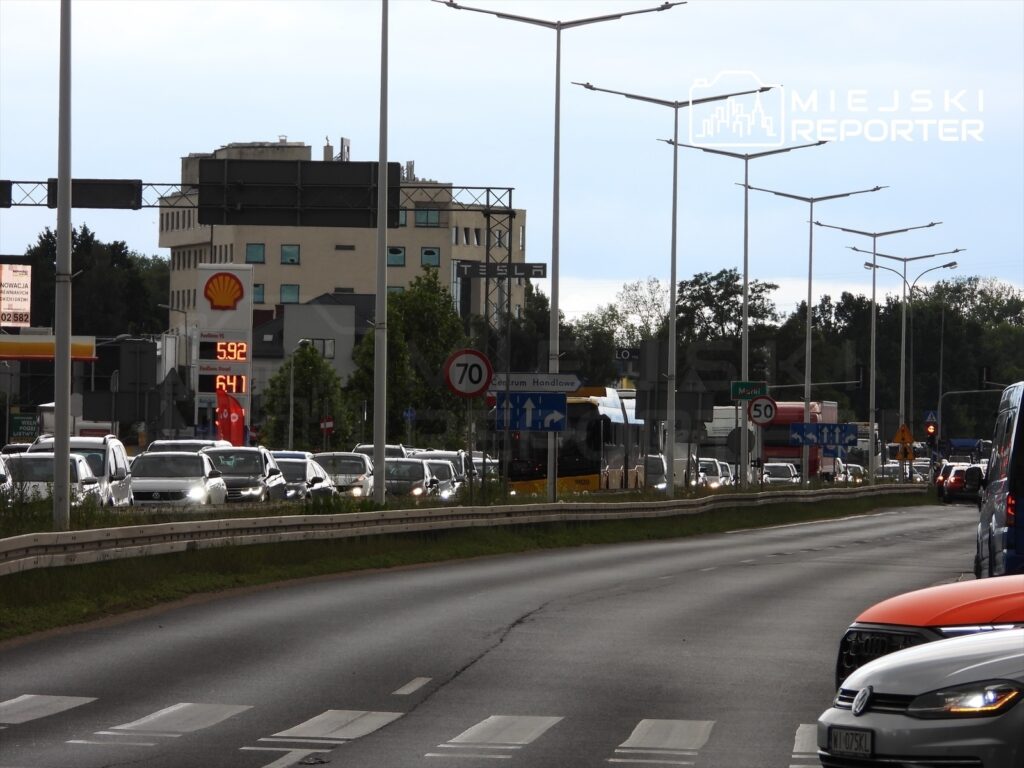 Ruchliwe skrzyżowanie z samochodami stojącymi w korku, stacja benzynowa Shell oraz widoczny wskaźnik cen paliw.