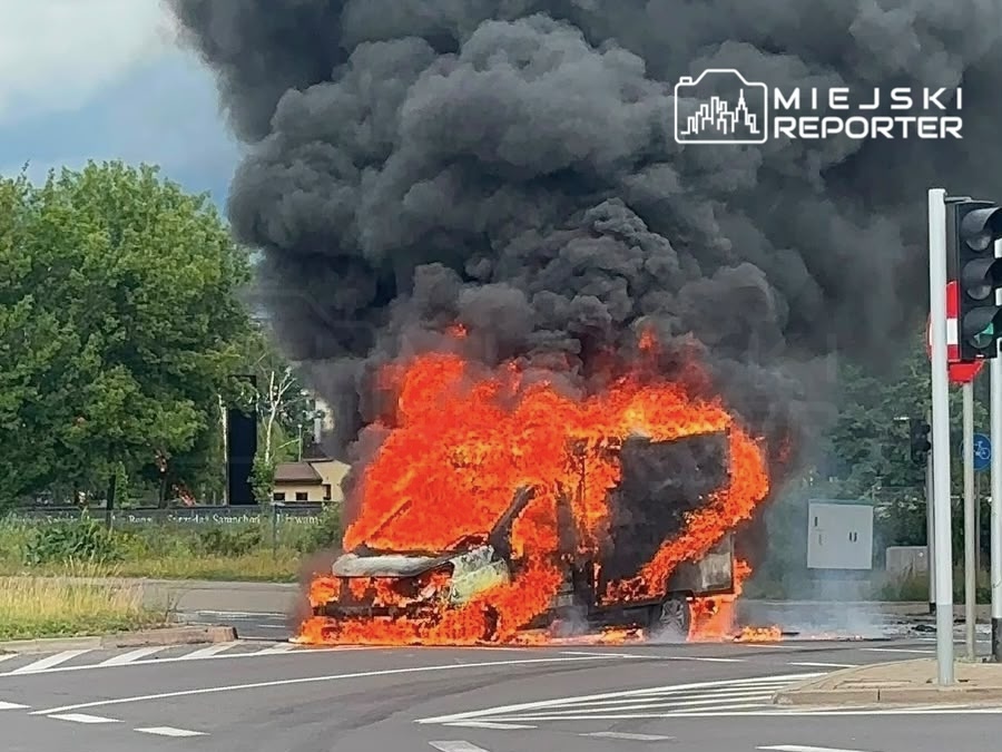 Samochód w ogniu na skrzyżowaniu, wydobywający gęsty czarny dym, w otoczeniu zieleni i sygnalizacji świetlnej.
