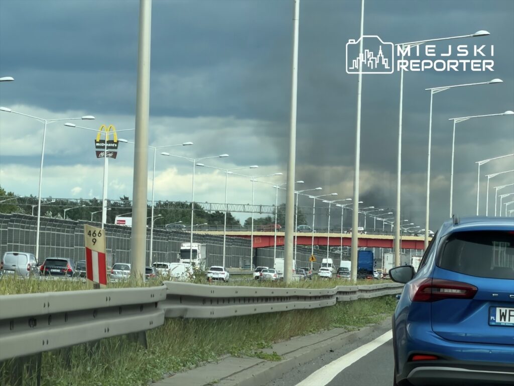 Na drodze widać samochody stojące w korku, a w tle unosi się ciemny dym nad budynkami. W górze widoczny znak McDonald's.