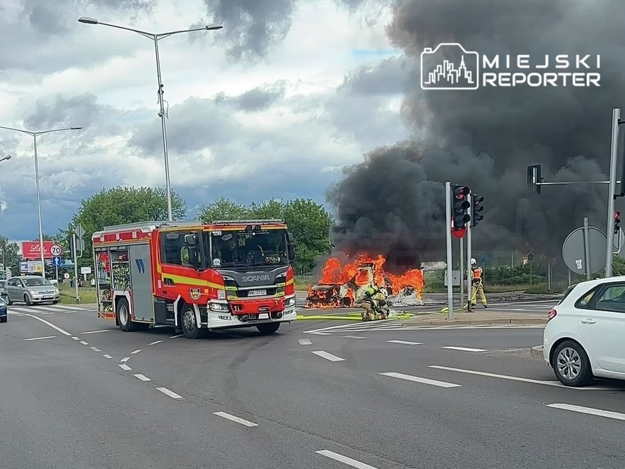 W tle gęsty czarny dym, strażacy gaszą płonący pojazd na skrzyżowaniu, obok przejeżdżające samochody.