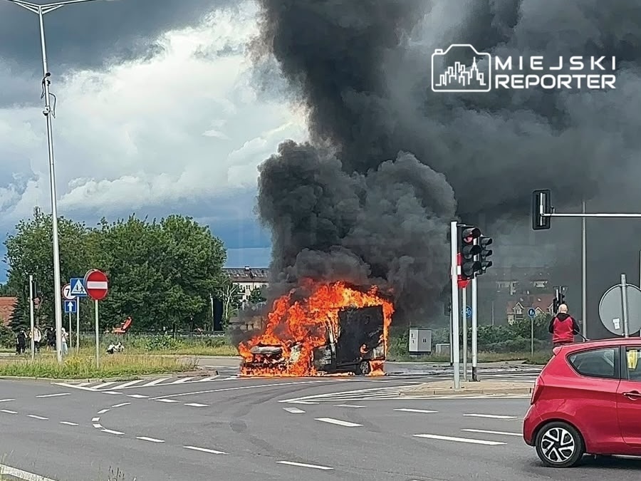 Pojazd płonący na skrzyżowaniu, wydobywający gęsty czarny dym, w tle widoczne znaki drogowe i chmury.