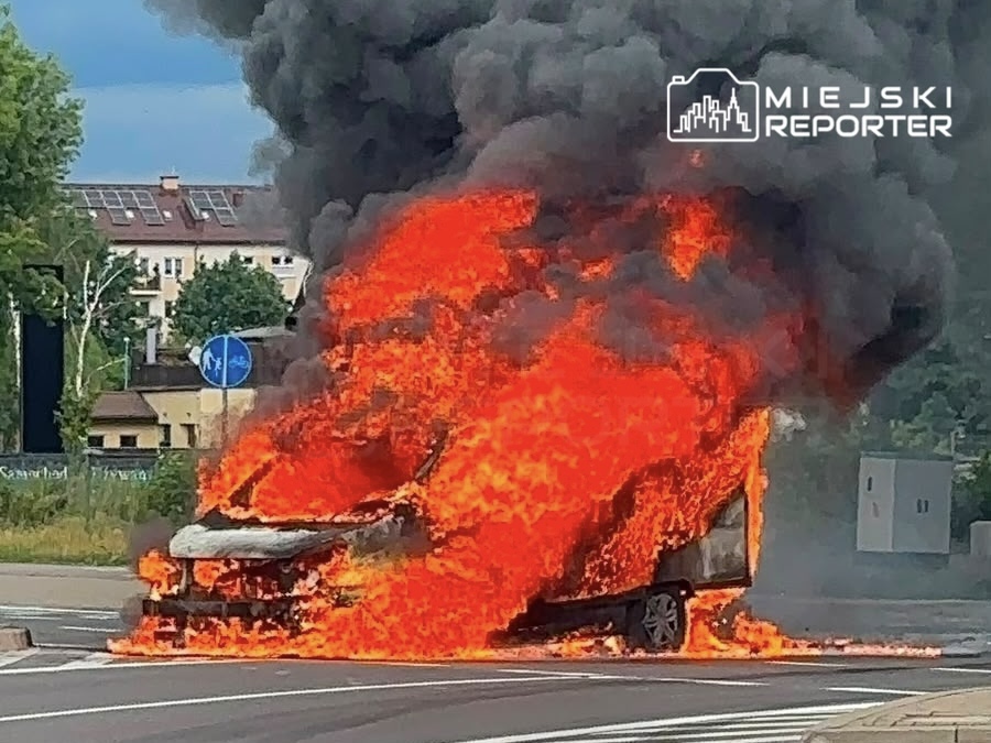 Samochód w płomieniach na skrzyżowaniu, emitujący gęsty czarny dym w tle.