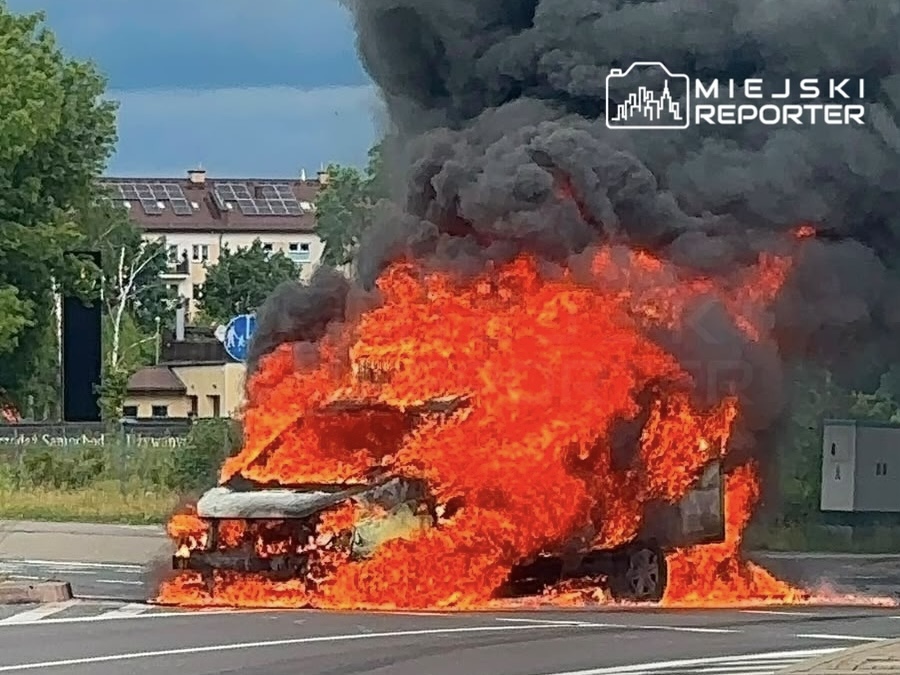 Samochód w płomieniach na skrzyżowaniu, wydobywający czarny dym, otoczony zielenią i budynkami w tle.