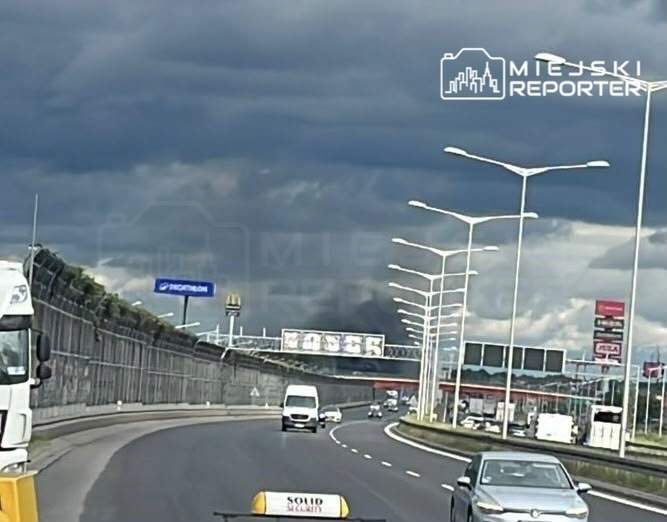 Ciemne chmury wiszą nad drogą, przy której widać samochody i oświetlenie uliczne. W tle billboardy reklamowe.