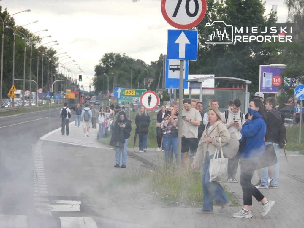 Grupa ludzi stoi na chodniku przy drodze, niektórzy korzystają z telefonów, w tle widać znaki drogowe i zieleń.