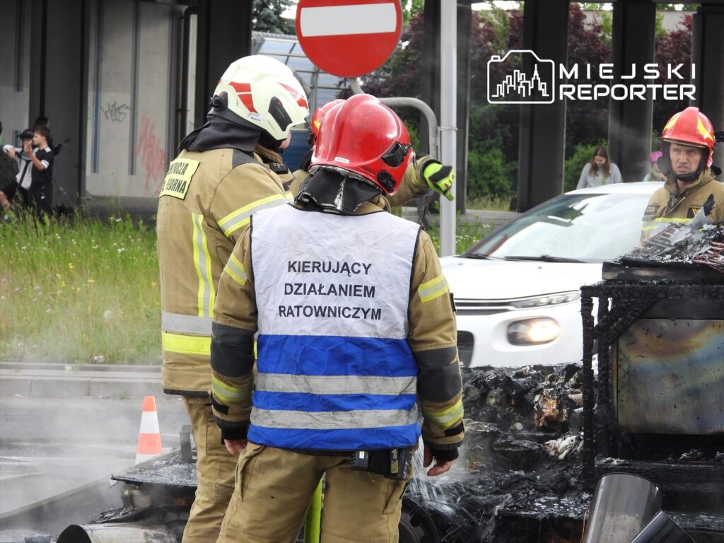 Grupa strażaków w hełmach i uniformach, monitorująca miejsce pożaru pojazdu na drodze, w tle przejeżdżające samochody.