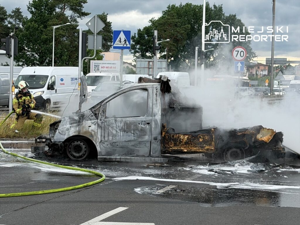 Strażak gaszący płonący pojazd na skrzyżowaniu, wokół widoczne inne samochody i znaki drogowe.