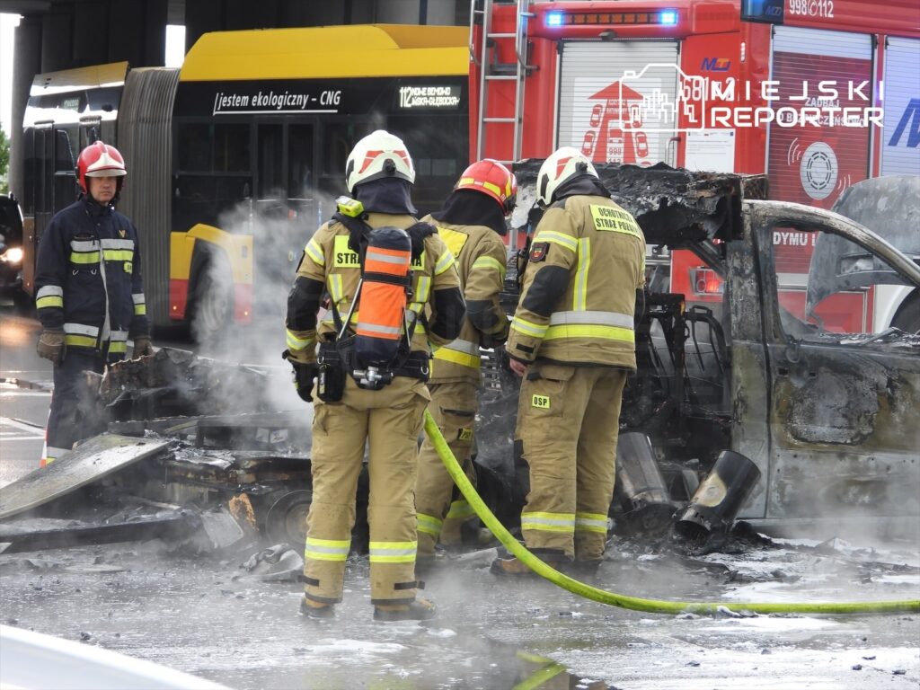 Grupa strażaków w mundurach gasząca płonący wrak samochodu na drodze, obok przejeżdżający autobus.