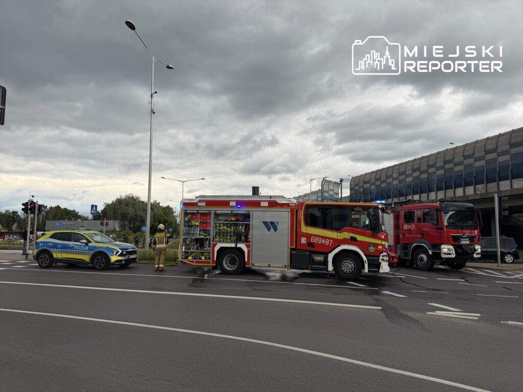Na skrzyżowaniu stoją wozu strażackie i radiowóz policyjny, w tle ciemne chmury.