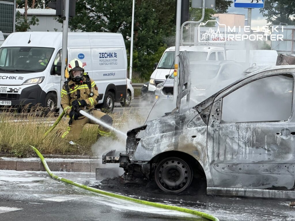 Strażak w hełmie gasi płonący samochód osobowy na drodze, obok stoją pojazdy dostawcze.