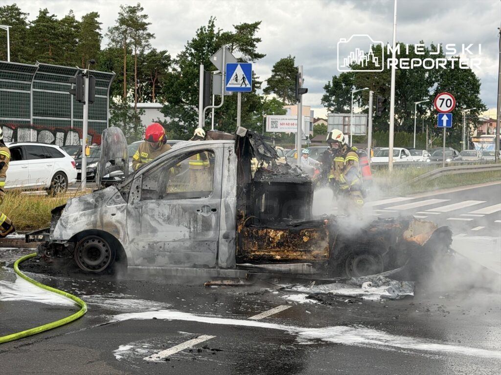 Strażacy gaszą płonący pojazd na skrzyżowaniu, wokół widoczne inne samochody i znaki drogowe.