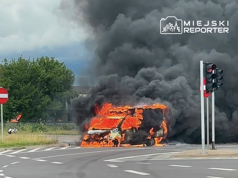 Pojazd w płomieniach na skrzyżowaniu, wokół gęsty dym i znaki drogowe.