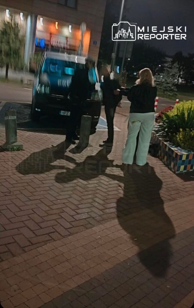 Grupa osób rozmawia obok czarnego samochodu w oświetlonym nocą otoczeniu parkingu.