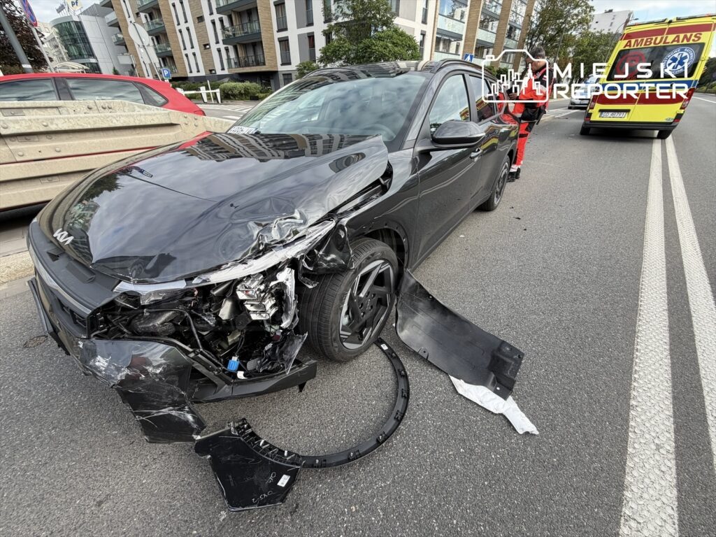 Czarny samochód osobowy z uszkodzoną przednią częścią stoi na jezdni, obok pracuje ratownik medyczny.