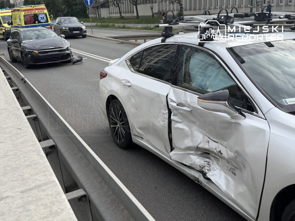 Dwa uszkodzone samochody na jezdni, z białym autem z wgniecioną stroną i czarnym pojazdem z uszkodzoną przednią częścią; w tle ambulans.