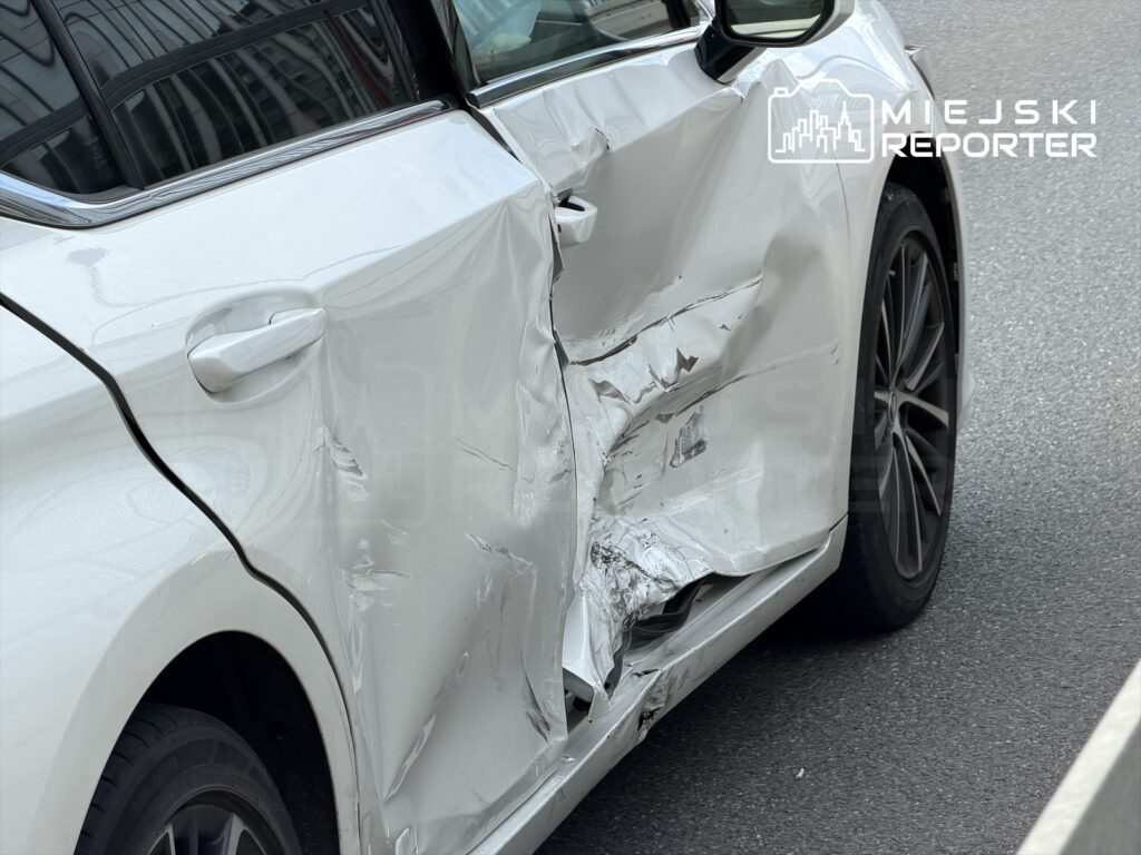 Uszkodzony biały samochód osobowy z wgniecioną boczną częścią, zaparkowany na ulicy.