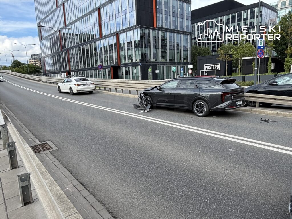 Czarny samochód osobowy z uszkodzeniami stoi na pasie drogi obok nowoczesnego biurowca, podczas gdy biały samochód przejeżdża obok.