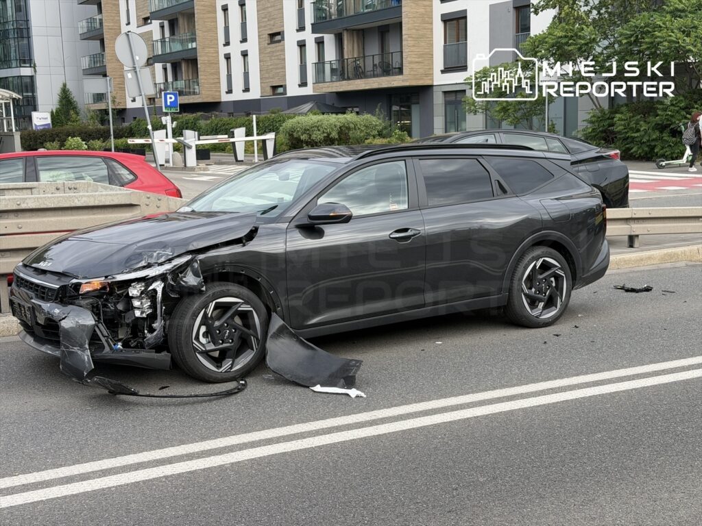 Czarny samochód osobowy z uszkodzoną przednią częścią stoi na ulicy obok nowoczesnych budynków mieszkalnych.