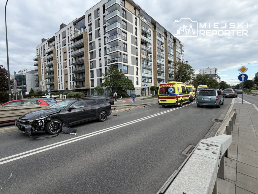 Czarny samochód osobowy z uszkodzeniami stoi na drodze, obok niego dwa ambulanse, w tle nowoczesny budynek mieszkalny.