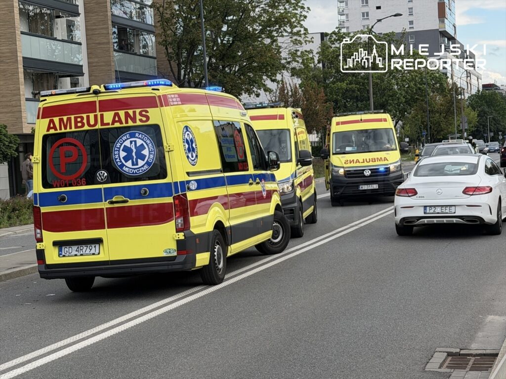Trzy karetki pogotowia z włączonymi sygnałami świetlnymi zatrzymane na ulicy, obok przejeżdżające samochody.
