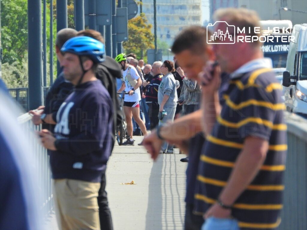 Grupa ludzi stojąca na moście, niektórzy z rowerami, w słoneczny dzień. W tle widać budynki.