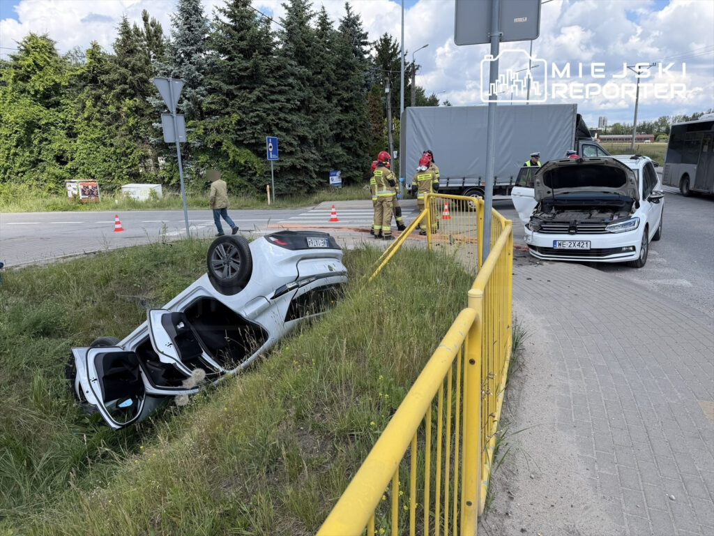 Samochód osobowy leży na dachu w rowie, a obok interweniują strażacy i policja. Na drodze widoczne pachołki.