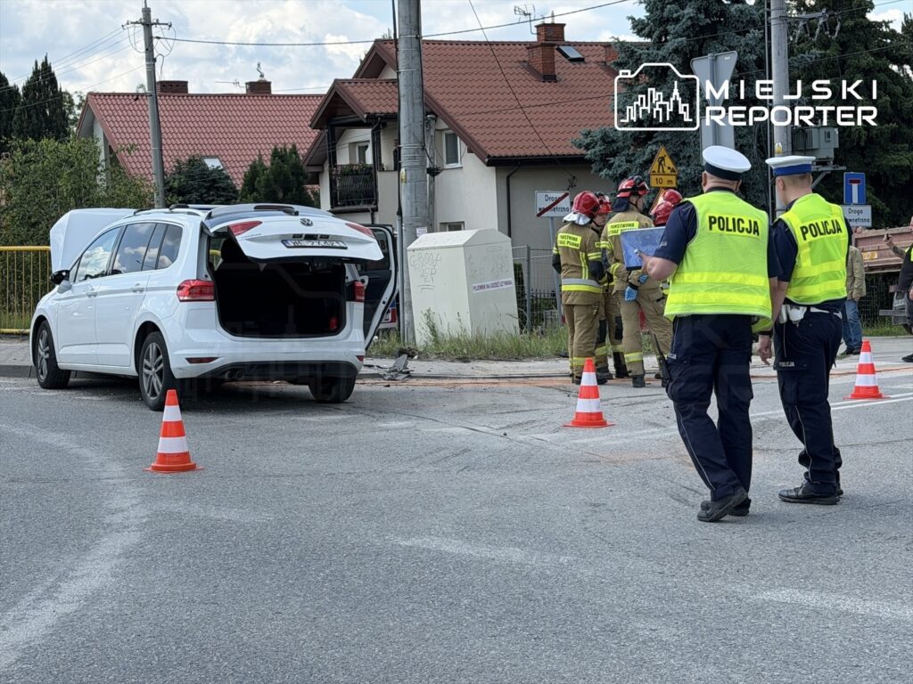 Biały samochód osobowy z otwartym bagażnikiem stoi na drodze, obok dwóch policjantów w kamizelkach odblaskowych oraz strażaków.