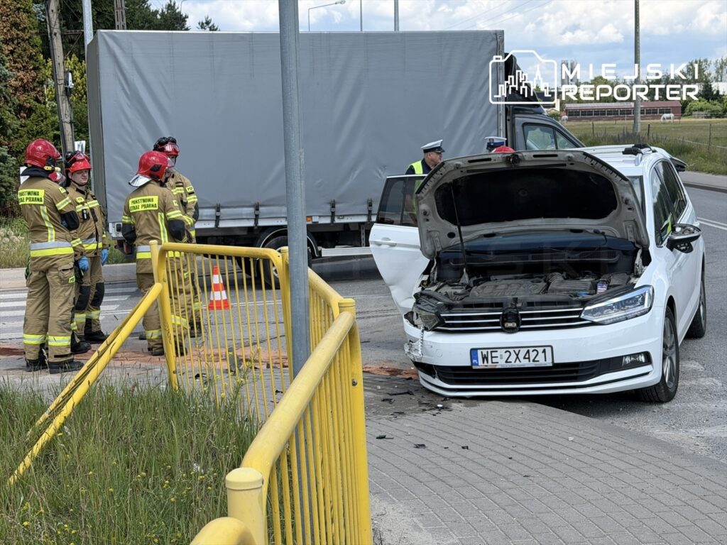 Grupa strażaków w mundurach z logo „Państwowa Straż Pożarna” ocenia uszkodzenia białego samochodu osobowego po kolizji.