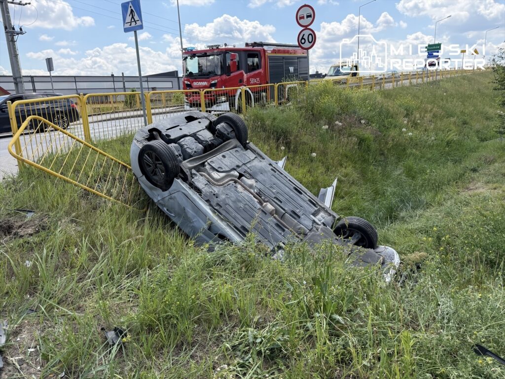 Samochód leżący na dachu w trawie obok drogi, obok widoczny wóz strażacki i znaki drogowe.