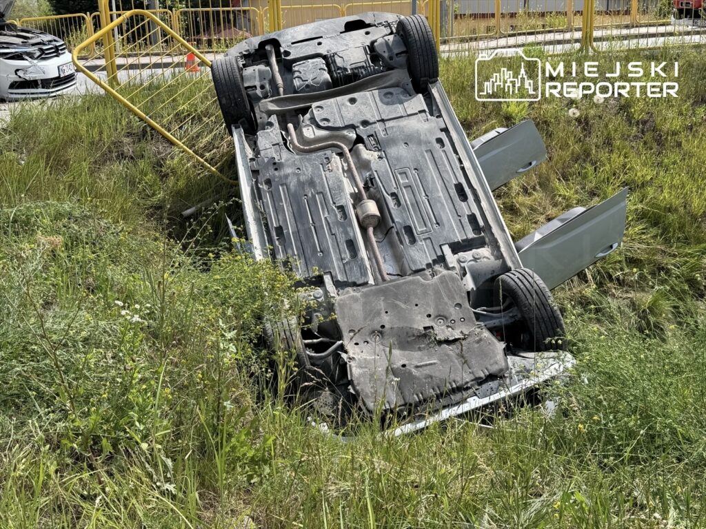 Samochód leżący na dachu w trawie obok drogi, z otwartymi drzwiami i widocznymi uszkodzeniami.
