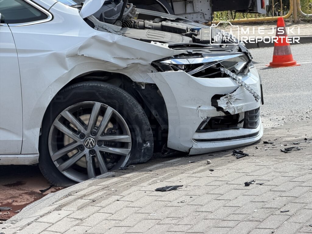 Uszkodzony biały samochód osobowy z zgniecionym przodem, stojący na krawężniku w miejskim otoczeniu.