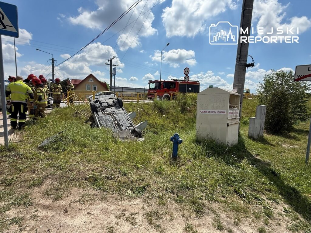 Grupa strażaków w mundurach obserwuje przewrócone auto na trawie obok drogi, w tle widoczny czerwony wóz strażacki.