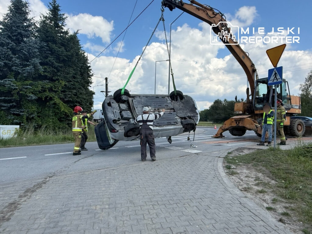 Grupa strażaków i pracowników w kamizelkach odblaskowych podnosi przewrócone auto za pomocą dźwigu na drodze.