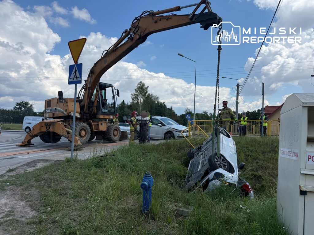 Na drodze stoi ciężki sprzęt budowlany, który podnosi przewrócony samochód z rowu, a obok pracują ratownicy.