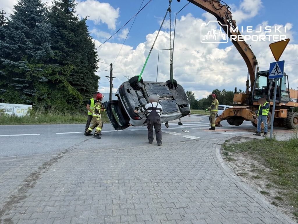Grupa strażaków podnosi przewrócone auto przy użyciu żurawia na skrzyżowaniu drogowym.