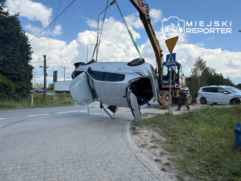 Samochód uniesiony przez dźwig przy drodze, z otwartymi drzwiami, obok pracownik w odblaskowej kamizelce.
