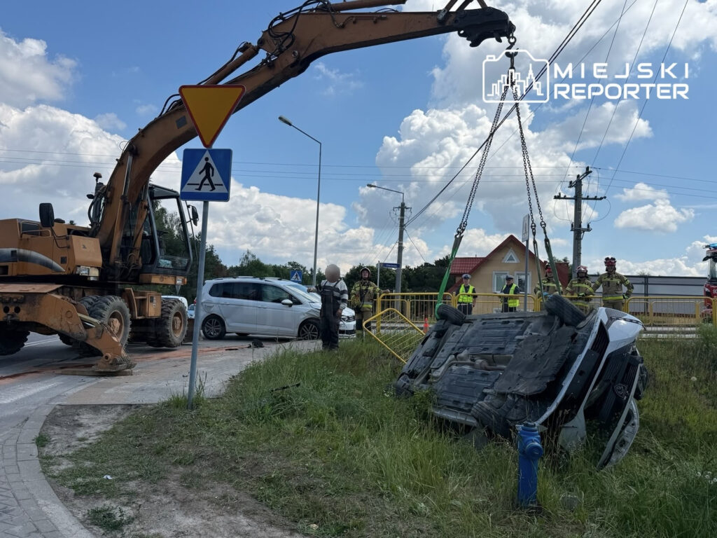 Wysoka koparka podnosi przewrócone auto na poboczu drogi, obok grupy strażaków i świadków zdarzenia.