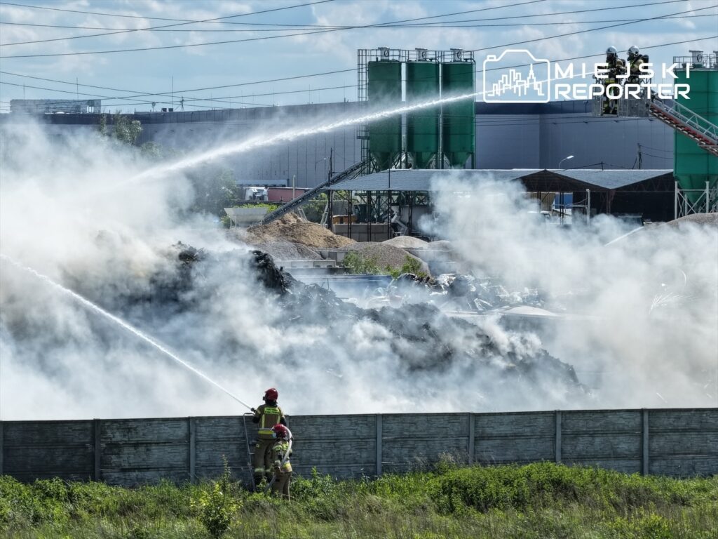 Strażacy gaszą pożar na wysypisku, używając wody z węży strażackich, otoczeni gęstym dymem.