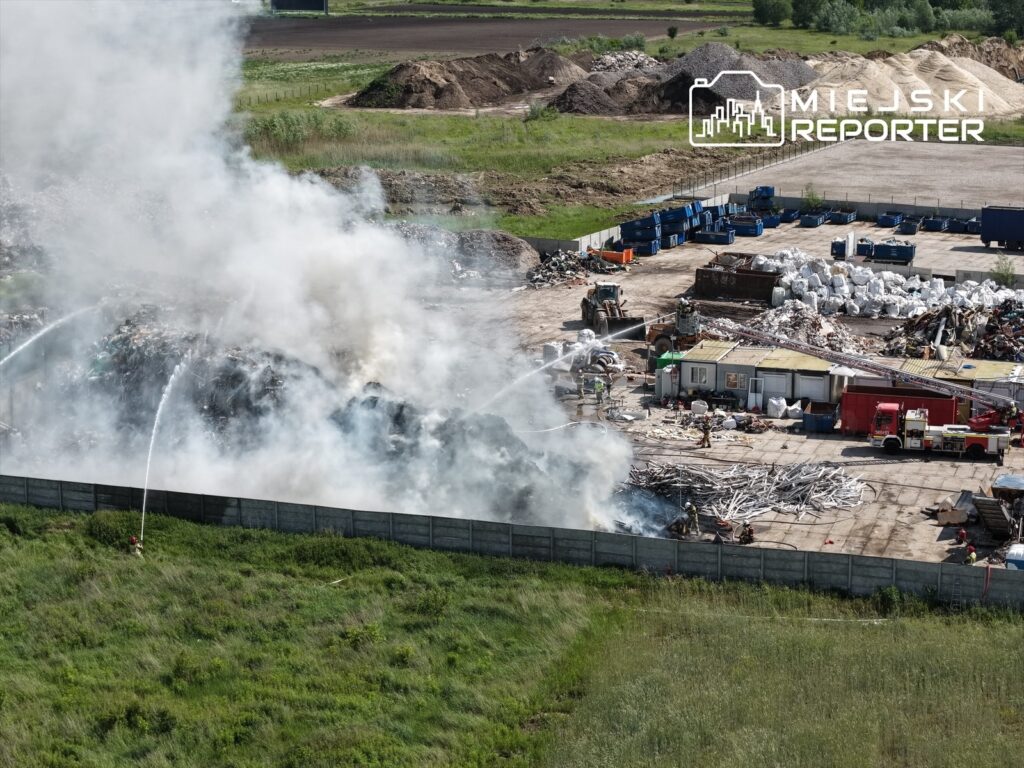 Ognisko pożaru na wysypisku odpadów, gdzie strażacy gaszą dymiące resztki z użyciem wody. W tle widoczne maszyny i kontenery.