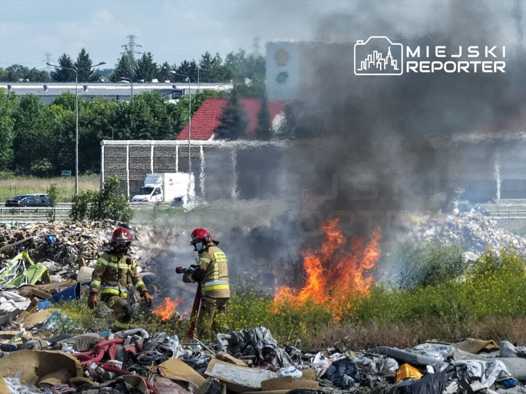 Dwaj strażacy gaszą pożar na wysypisku śmieci, w tle widać dym i płomienie oraz samochód dostawczy.