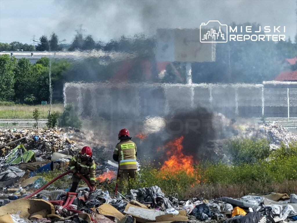 Strażacy w mundurach gaszą pożar na wysypisku śmieci, otoczeni dymem i płomieniami.