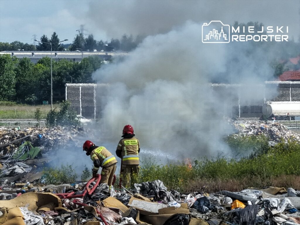 Dwaj strażacy w hełmach gaszą pożar na wysypisku śmieci, otoczeni chmurami dymu i odpadami.