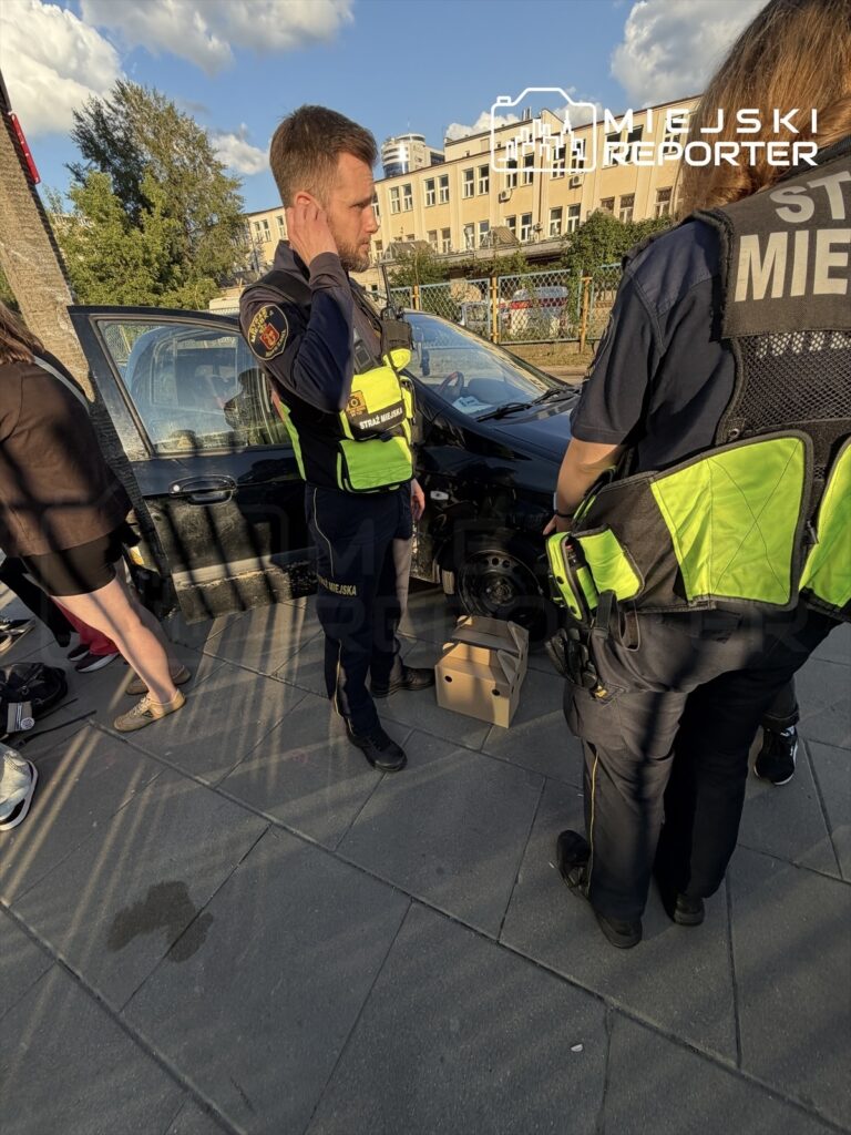 Mężczyzna w mundurze straży miejskiej rozmawia z kolegą, obok uszkodzonego samochodu na chodniku.