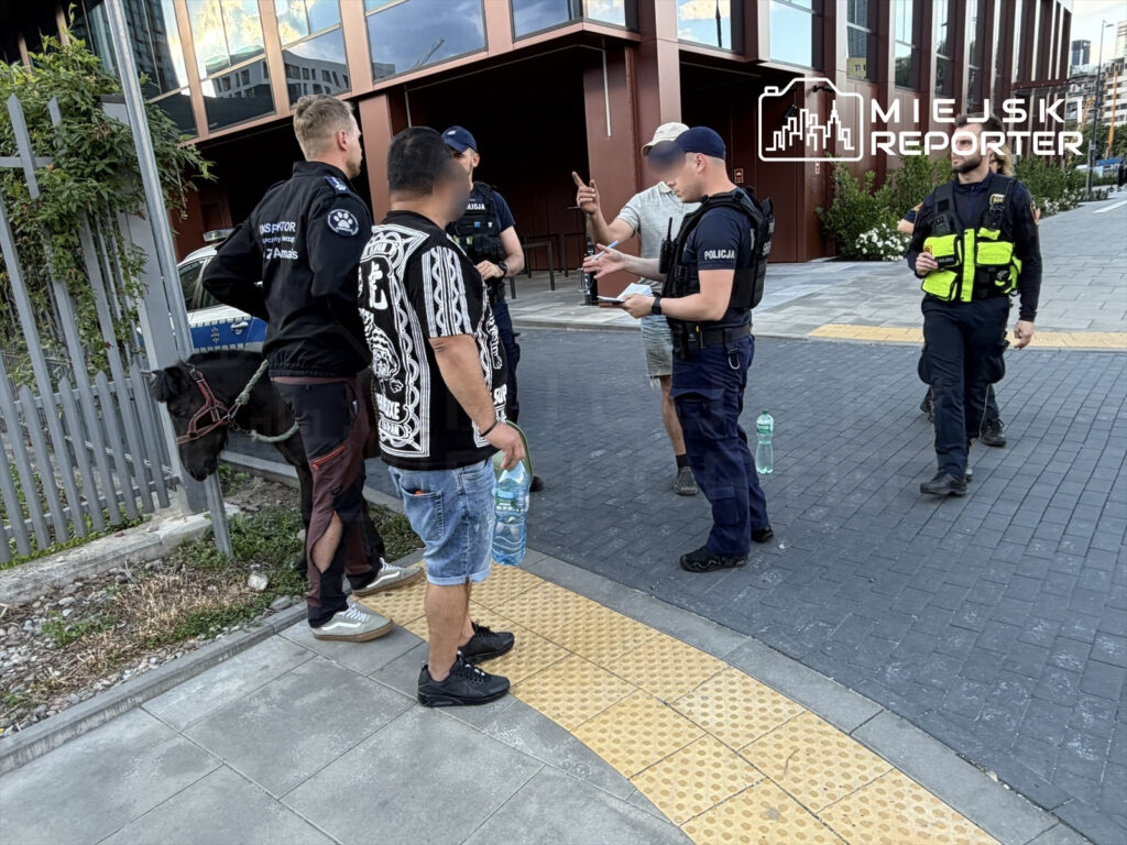 Czterech funkcjonariuszy policji rozmawia z mężczyznami na chodniku obok czarnego konia przypiętego do ogrodzenia.