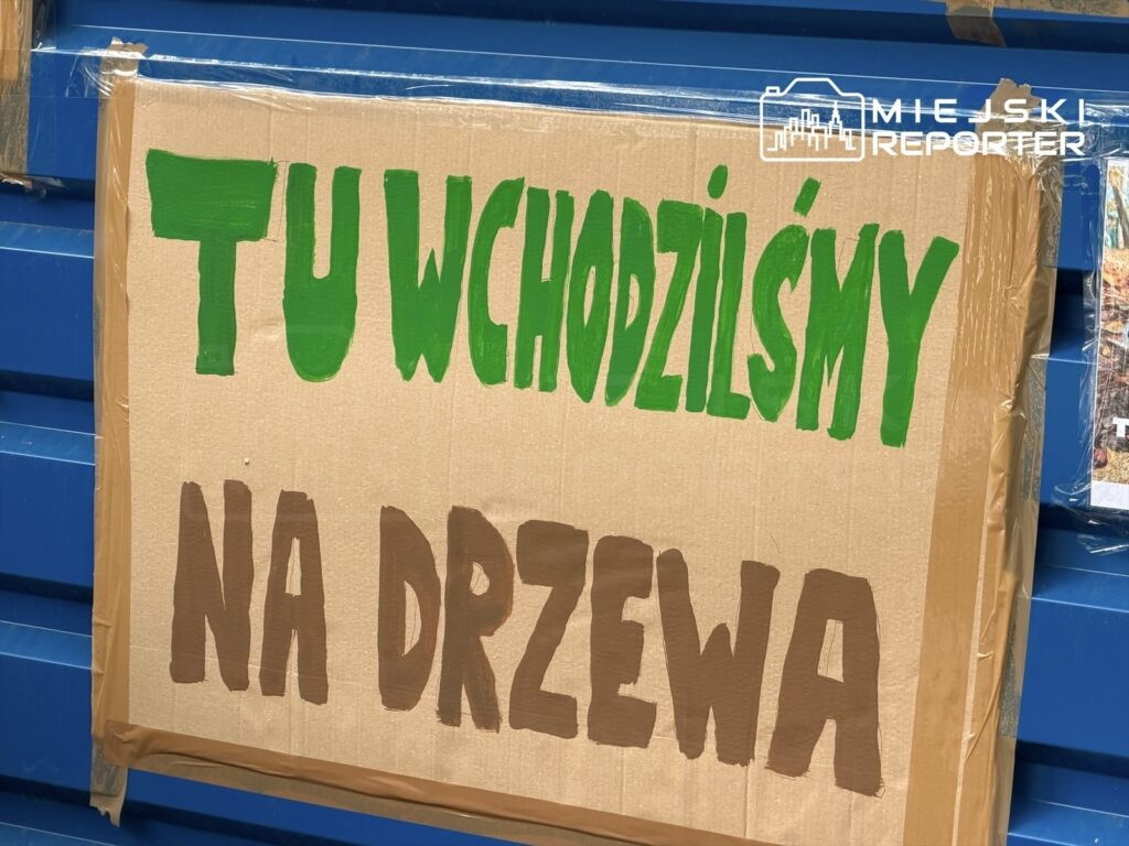 Na tekturowym plakacie widnieje napis „TU WCHODZILIŚMY NA DRZEWA” w zielonych i brązowych literach. Plakat przymocowany jest do niebieskiej ściany.