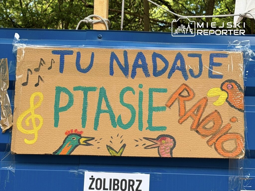Kolorowy banner z napisem "TU NADAAJE PTASIE RADIO" ozdobiony rysunkami ptaków i nutami, zawieszony w zielonym otoczeniu.