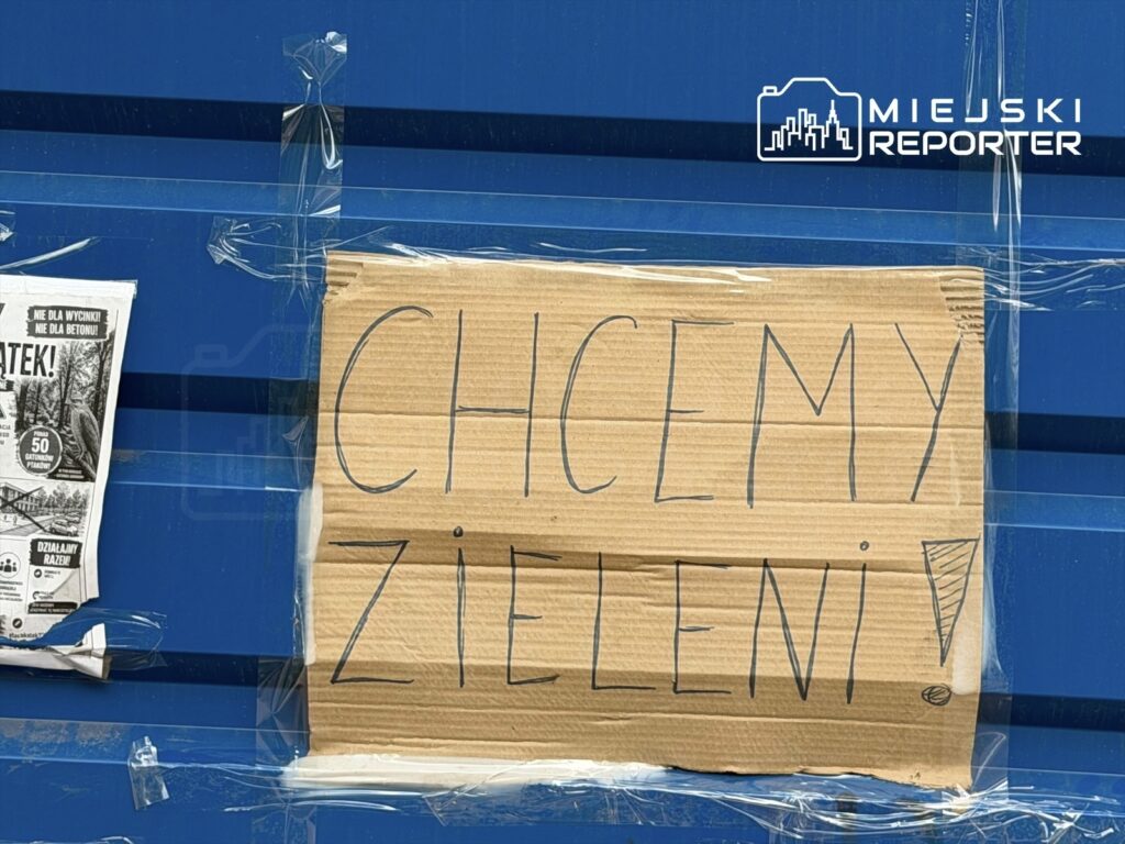 Na niebieskiej ścianie umieszczona tektura z napisem "CHCEMY ZIELENI!" przyklejona taśmą. Obok widoczny plakat.